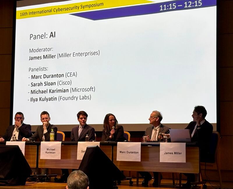 Panelists seated on stage beneath the projected AI panel programme at the 16th International Cybersecurity Symposium.